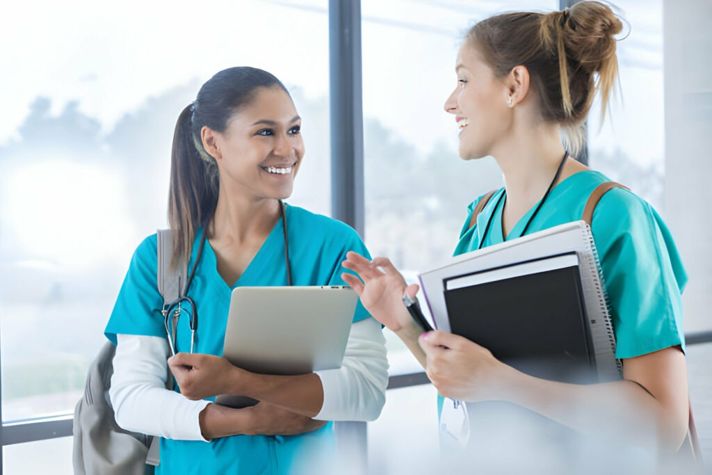 Two healthcare professionals in teal scrubs engaging in a discussion with a laptop and notebooks