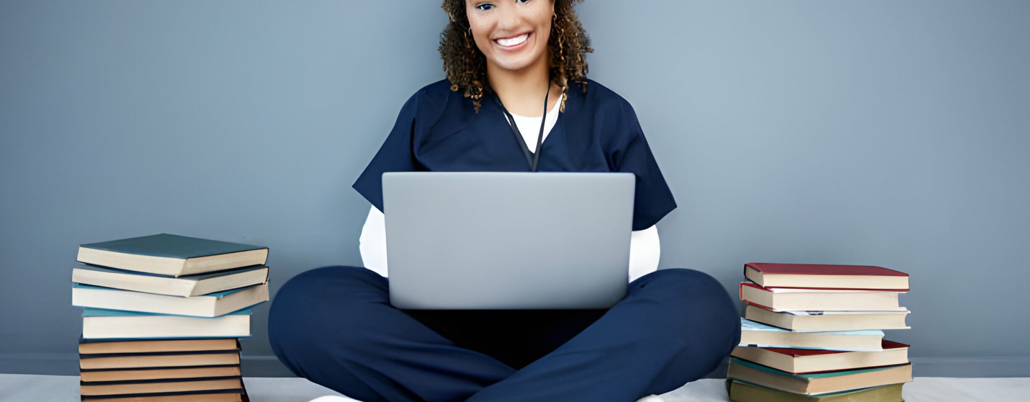 A healthcare professional smiling, working on a laptop with stacks of books beside her
